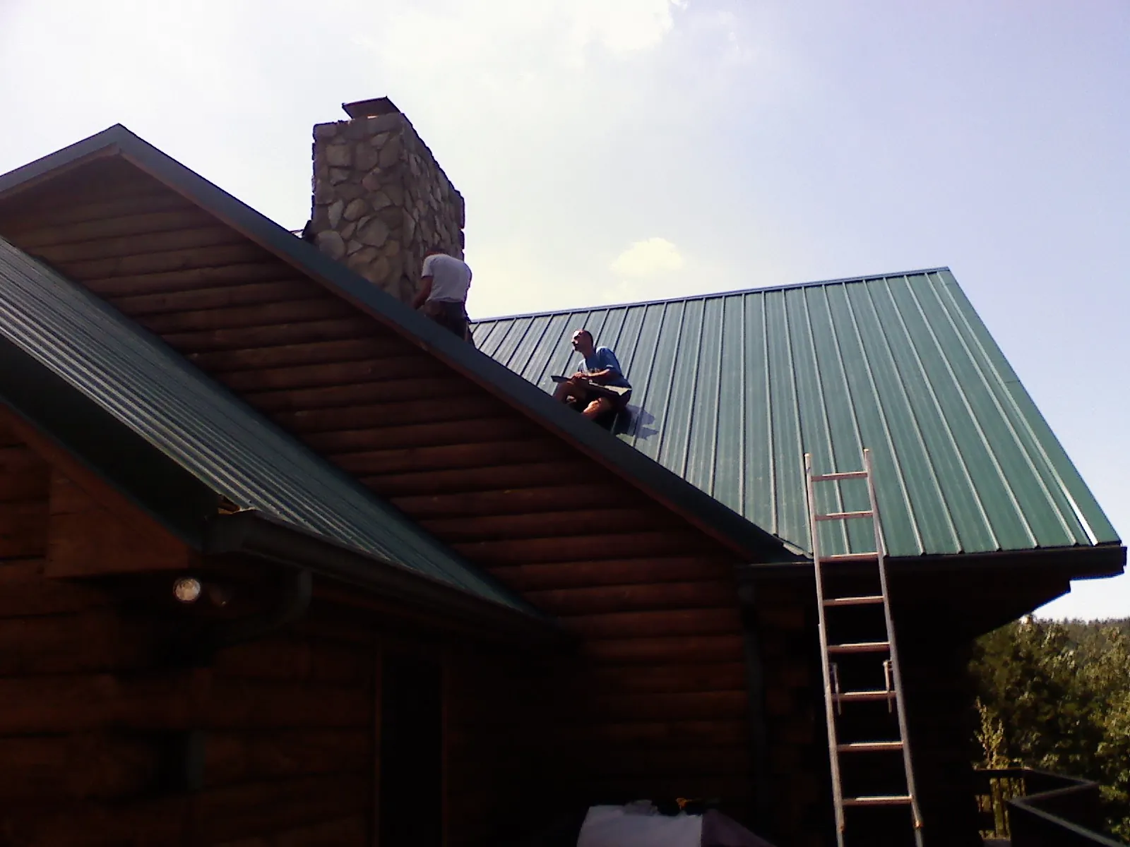 Roofers installing green metal panels beside a stone chimney on a log home in Union City